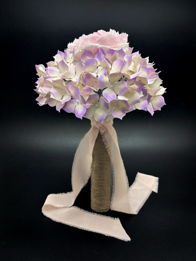 Profilo intero di un bouquet di carta con ortensie viola, impugnatura rivestita in corda naturale e un lungo fiocco in chiffon rosa antico.