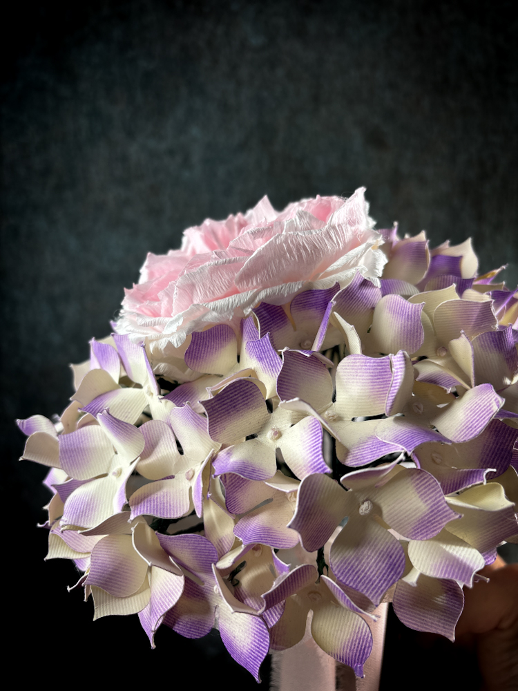 Vista dall'alto di un bouquet sposa con ortensie di carta rigida sfumata lilla e crema che circondano una rosa inglese rosa confetto.